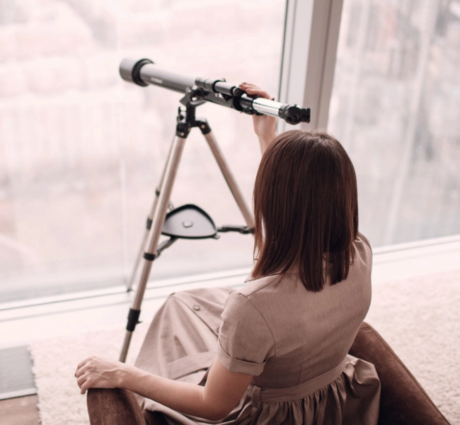 A woman uses a telescope in an office setting to look out a window.