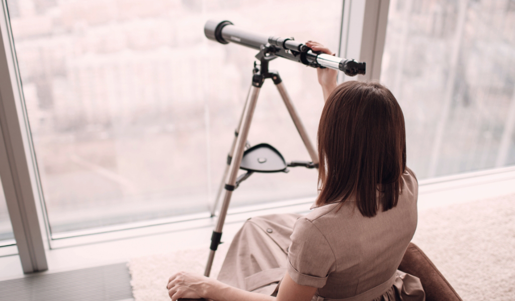 A woman uses a telescope to look out her window.