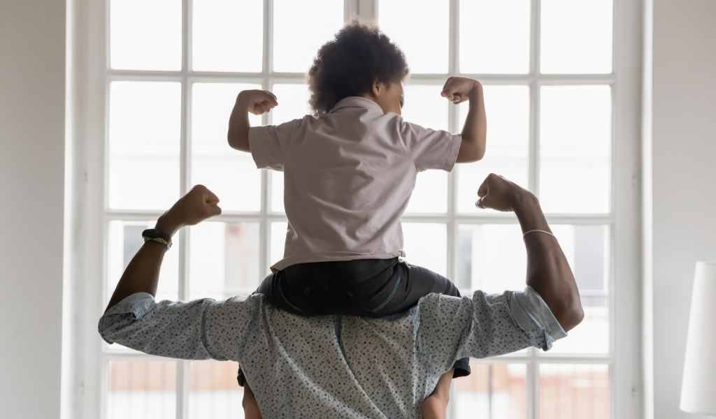 A man carries his son on his shoulders, while both of them flex their muscles.